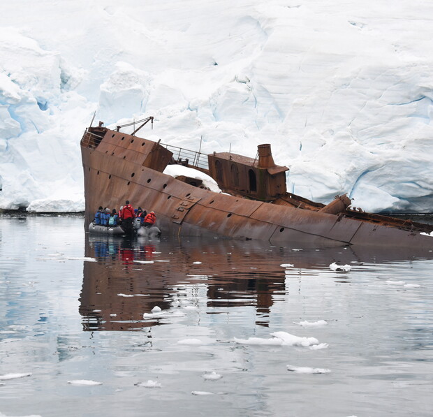 A rusted shipwreck partially submerged in icy water, with people in an inflatable boat inspecting it, surrounded by large ice formations in a cold, remote environment.